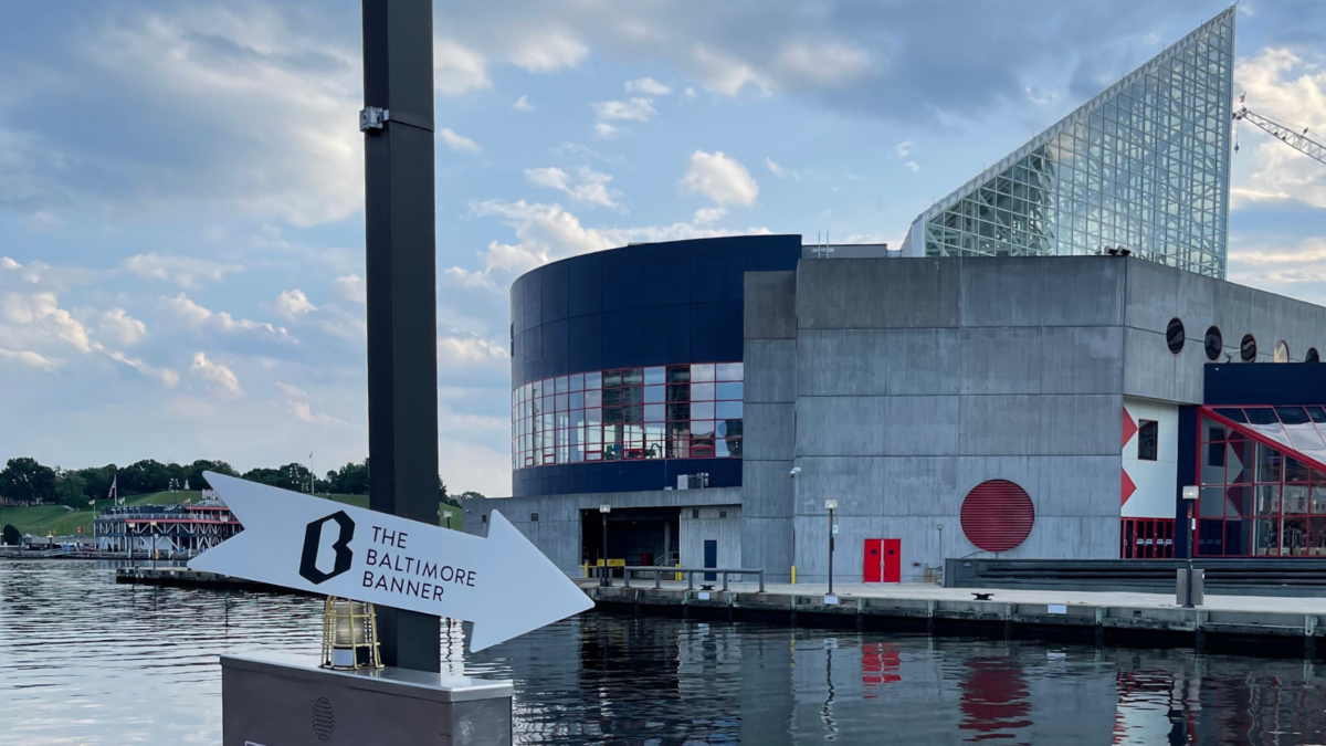 An arrow-shaped reads "The Baltimore Banner." The National Aquarium in Baltimore is in the background.