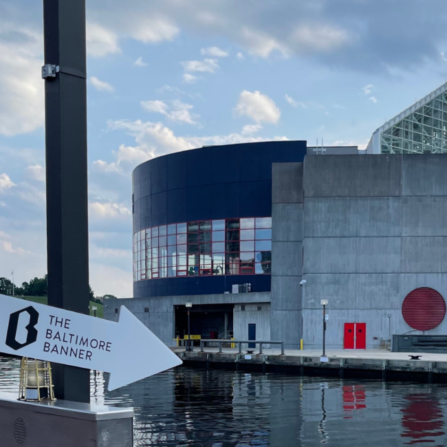 An arrow-shaped reads "The Baltimore Banner." The National Aquarium in Baltimore is in the background.