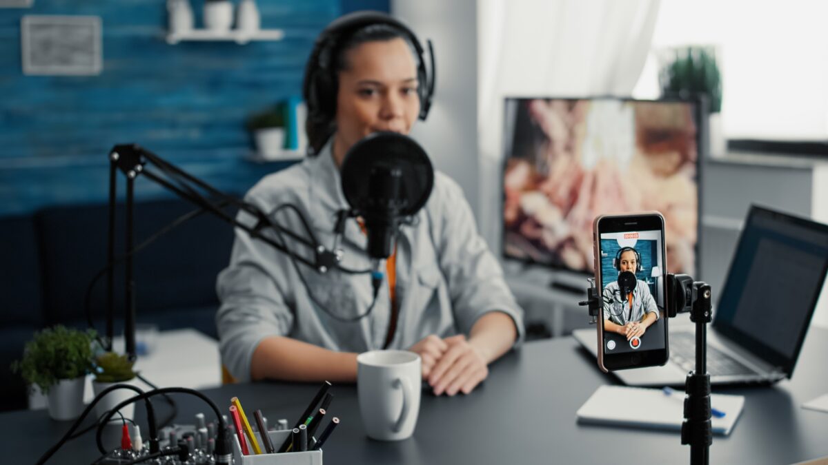 A woman in a blue shirt is sitting at a desk recording herself on a smartphone with a microphone
