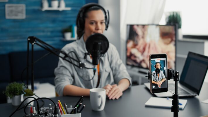 A woman in a blue shirt is sitting at a desk recording herself on a smartphone with a microphone